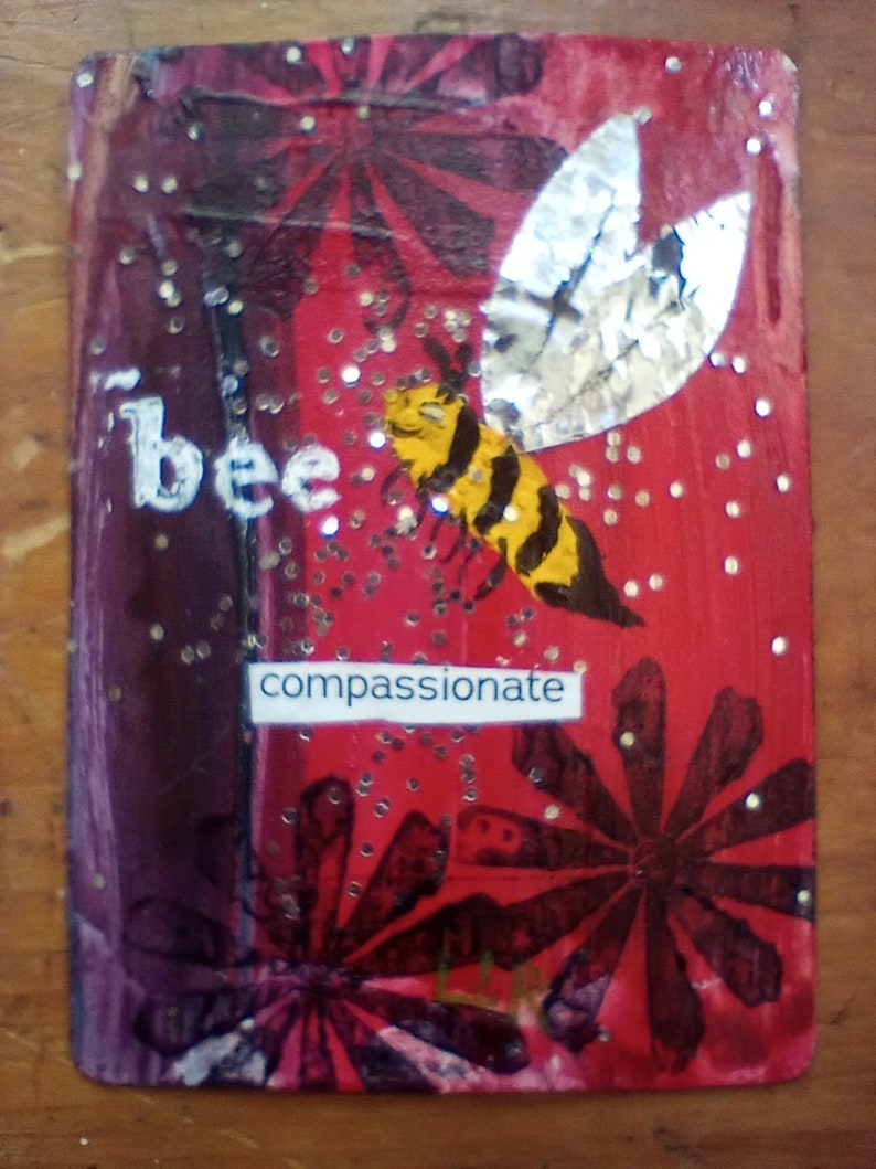 May include: A rectangular artwork with a red and purple background, featuring a yellow and black bee with silver wings. The word "bee" is written in white, and the word "compassionate" is on a white label. Glitter is scattered across the surface.