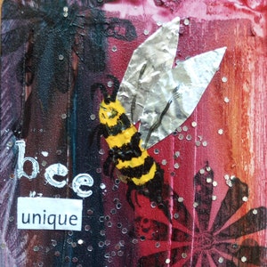 May include: A mixed media artwork featuring a bee with yellow and black stripes and metallic wings. The background is a blend of red, black, and orange hues with scattered glitter. The words "bee unique" are displayed in white.