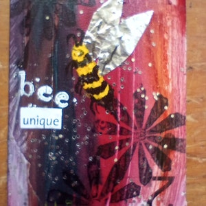 May include: A rectangular artwork with a bee illustration and the words "bee unique." The bee has yellow and black stripes with silver foil wings. The background transitions from dark purple to red with floral patterns and glitter.