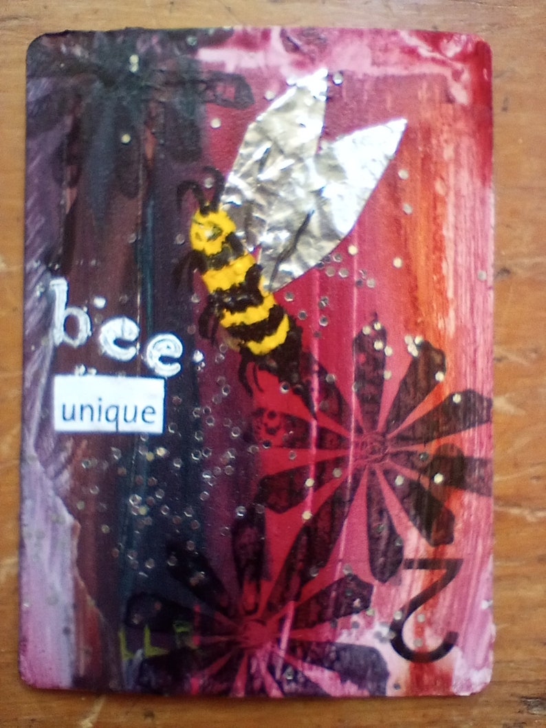 May include: A rectangular artwork with a bee illustration and the words "bee unique." The bee has yellow and black stripes with silver foil wings. The background transitions from dark purple to red with floral patterns and glitter.