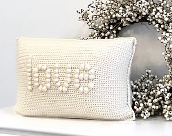 Op de afbeelding: Crèmekleurig rechthoekig gehaakt kussen met het woord "LOVE" in reliëf bobbelsteek. Het kussen ligt op een wit oppervlak, met een witte bessenkrans op de achtergrond. Een decoratief woonaccessoire.