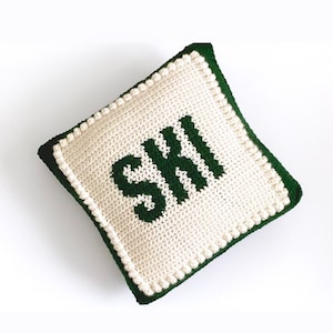 May include: A white crocheted cushion with a green embroidered word "SKI" on the front. The cushion has a green border.