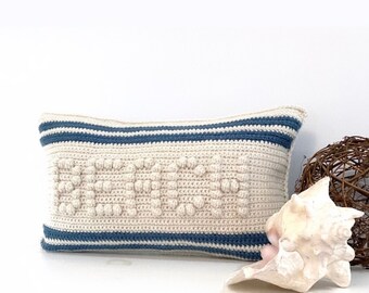 May include: A cream-colored crocheted pillow with blue stripes and a textured design. The pillow is sitting on a white surface with a seashell and a wicker ball.