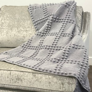 May include: A light gray crocheted blanket with a textured, raised dot pattern draped over a gray sofa. The blanket's design features a grid-like arrangement of bobbles, creating a cozy and tactile appearance. The blanket is made of soft yarn.