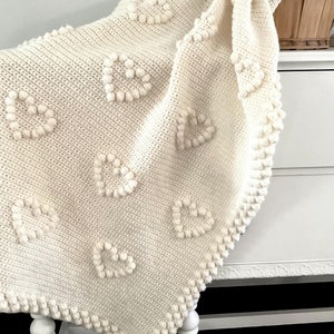 May include: A cream-colored crocheted blanket with a heart pattern and a textured border. The blanket is draped over a white dresser with a basket on top.