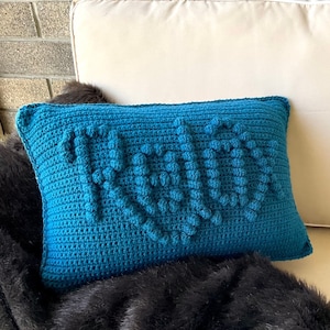 Puede incluir: Una almohada de punto de crochet azul turquesa con un diseño texturizado. La almohada tiene un patrón en relieve que se asemeja a la palabra "RAD".