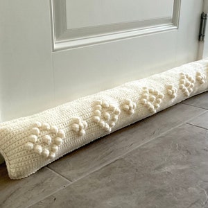 May include: A white crocheted doorstop with a heart pattern and raised bobbles. The doorstop is lying on a tiled floor.