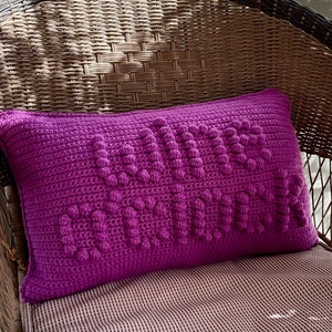 Puede incluir: Una almohada de crochet morado con el texto "Love is Love" en letras en relieve.