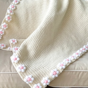 May include: A crocheted baby blanket in cream with a border of white and pink flowers.