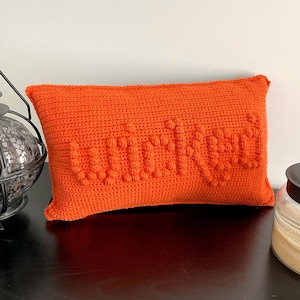 May include: Orange crocheted pillow with the word "Cozy" spelled out in raised stitches.
