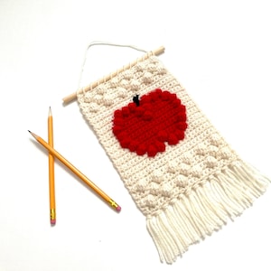 May include: A white crocheted wall hanging with a red apple design. The apple has a black stem and leaf. The hanging has white fringe on the bottom.