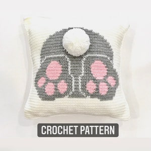 May include: Crochet pattern for a gray bunny pillow with pink paw prints and a white pom-pom tail.  "CROCHET PATTERN" is written below the pillow.