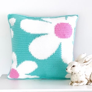 May include: A turquoise blue cushion with a white crocheted daisy design and pink centres. The cushion is sitting on a white shelf with a white ceramic rabbit figurine.