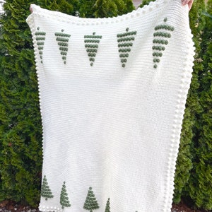 May include: A white crocheted blanket with a green embroidered design of Christmas trees. The blanket has a white crocheted border with small white pom-poms.