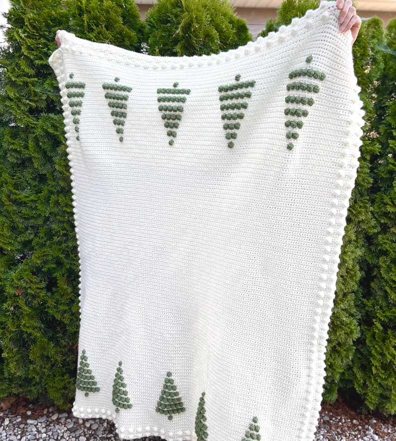 May include: A white crocheted blanket with a green embroidered design of Christmas trees. The blanket has a white crocheted border with small white pom-poms.