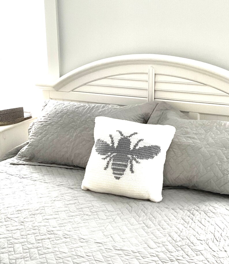 May include: A white pillow with a gray crocheted bee design on a bed with gray bedding.