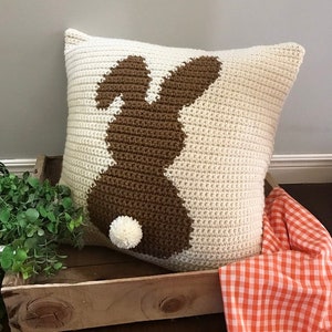 May include: A crocheted pillow with a brown bunny silhouette and a white pom-pom tail. The pillow is on a wooden crate with a red and white checkered fabric.