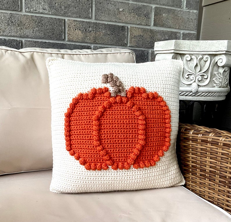 May include: A square cream-colored crochet pillow featuring a large, textured orange pumpkin design in the center. The pumpkin is crafted with raised bobble stitches along its edges and segments, topped with a small, light brown crocheted stem.