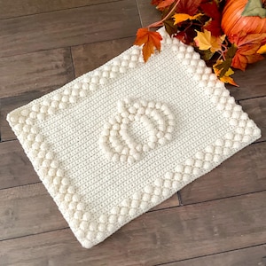May include: Cream-colored, rectangular crochet blanket with a pumpkin design. The blanket features a textured border and a pumpkin shape in the center. Perfect for fall decor, this handmade blanket adds a cozy touch to any space.