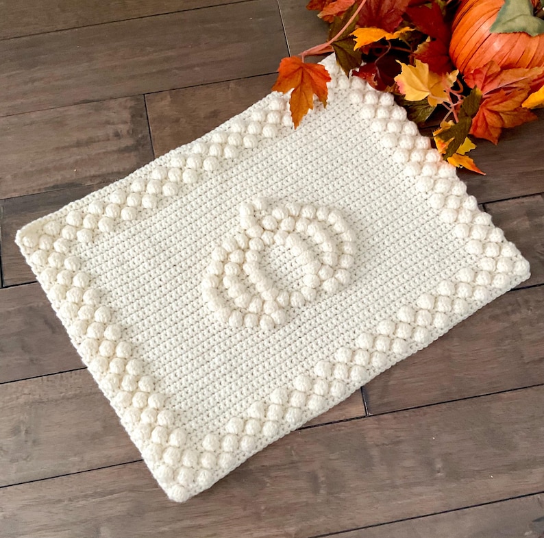 May include: Cream-colored, rectangular crochet blanket with a pumpkin design. The blanket features a textured border and a pumpkin shape in the center. Perfect for fall decor, this handmade blanket adds a cozy touch to any space.