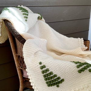 May include: A white crocheted throw blanket with a green pom-pom design. The blanket is draped over a wicker chair.