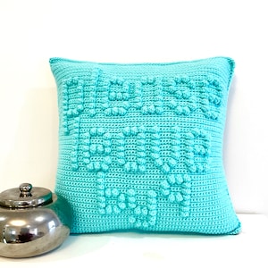 Puede incluir: Una almohada de punto de crochet azul turquesa con un patrón texturizado. La almohada es de forma cuadrada y tiene una textura suave y esponjosa.