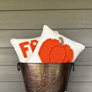 May include: Two square crochet pillows with off-white backgrounds are nestled in a dark bronze metal bucket with decorative scrollwork. One pillow features a textured orange pumpkin, while the other displays the partial orange text FR.