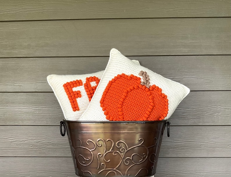 May include: Two square crochet pillows with off-white backgrounds are nestled in a dark bronze metal bucket with decorative scrollwork. One pillow features a textured orange pumpkin, while the other displays the partial orange text FR.