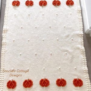 May include: Cream-colored, hand-crocheted blanket with orange pumpkin appliqu&eacute;s. The blanket features a textured bobble stitch and a border of small, round bobbles. The text "Seacliffe Cottage Designs" is visible on the blanket.