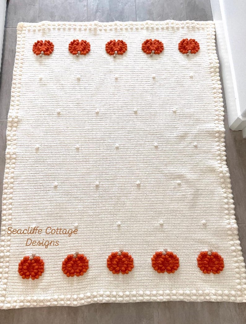 May include: Cream-colored, hand-crocheted blanket with orange pumpkin appliqu&eacute;s. The blanket features a textured bobble stitch and a border of small, round bobbles. The text "Seacliffe Cottage Designs" is visible on the blanket.