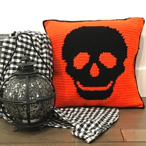 May include: Orange and black crocheted cushion with a skull design. The cushion is on a black and white checked fabric.