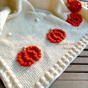 May include: Cream-colored crocheted blanket with orange pumpkin appliqu&eacute;s. The blanket features a textured stitch pattern and a border of bobble stitches. The pumpkins are detailed with brown stems. "Seacliff Cottage Designs" is printed on the blanket.
