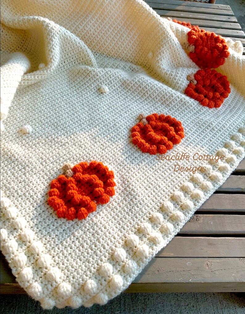 May include: Cream-colored crocheted blanket with orange pumpkin appliqu&eacute;s. The blanket features a textured stitch pattern and a border of bobble stitches. The pumpkins are detailed with brown stems. "Seacliff Cottage Designs" is printed on the blanket.