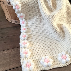 May include: A crocheted baby blanket in a cream color with a border of white and pink flowers.