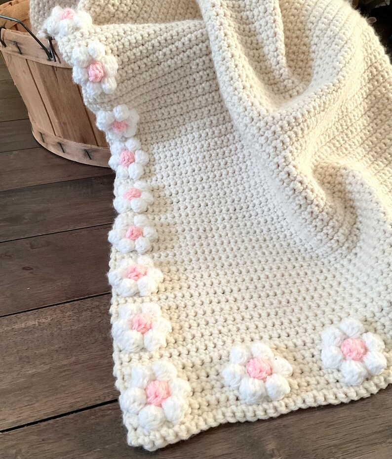 May include: A crocheted baby blanket in a cream color with a border of white and pink flowers.
