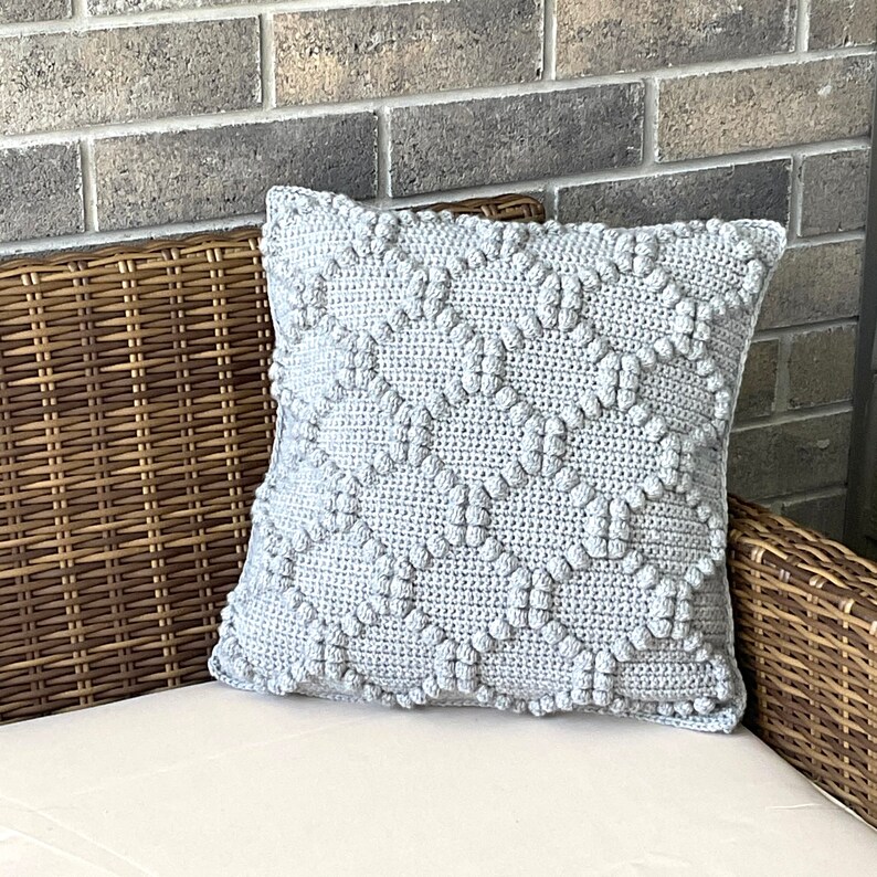 May include: A gray crocheted pillow with a textured diamond pattern. The pillow is sitting on a wicker chair.