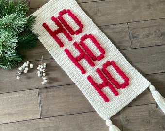 May include: White crocheted wall hanging with red pom-poms spelling out "HO HO HO". The hanging has two white tassels.