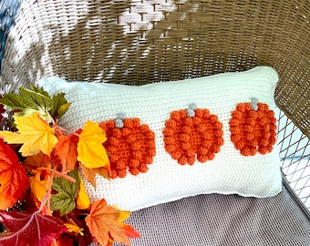 Puede incluir: Una almohada blanca de crochet con tres calabazas naranjas de crochet. La almohada está sentada en una silla de mimbre con hojas de otoño en primer plano.