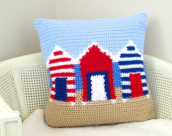 May include: A crocheted pillow with a beach scene featuring three colorful beach houses with red, white, and blue stripes. The houses are on a tan sand background with a light blue sky.