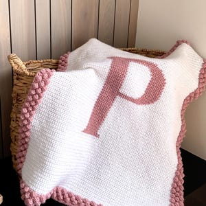 May include: A white crocheted blanket featuring a large, centered dusty pink letter P. The blanket is finished with a textured bobble stitch border in the same dusty pink yarn, creating a distinct edge around the rectangular white fabric.