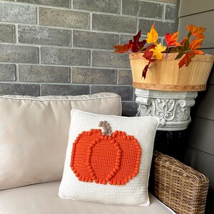May include: A square off-white crochet pillow featuring a centered orange pumpkin design with a brown stem. The pumpkin is crafted with a textured stitch pattern, and the pillow has a clean, simple border. The design is flat and centered on the front.