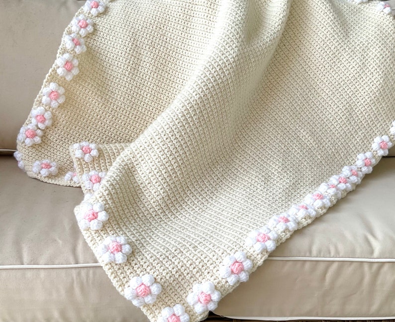 May include: A crocheted baby blanket in cream with a border of white and pink flowers.
