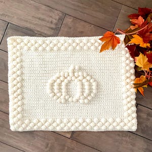 May include: Cream-colored, rectangular crochet pumpkin mat with a textured border. The pumpkin design is in the center of the mat. Autumn leaves are in the upper right corner. The mat is on a wooden surface.