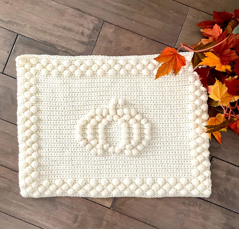 May include: Cream-colored, rectangular crochet pumpkin mat with a textured border. The pumpkin design is in the center of the mat. Autumn leaves are in the upper right corner. The mat is on a wooden surface.