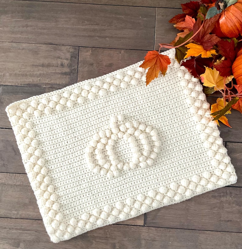 May include: Cream-colored, rectangular crochet blanket with a pumpkin design in the center. The blanket has a textured border and is made of soft yarn. Autumn leaves and pumpkins are in the background, suggesting a fall theme.