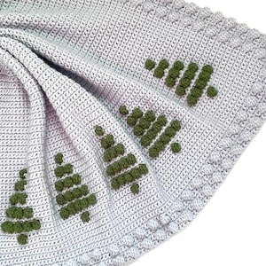 Pode incluir: Uma manta de crochê cinza com um design de árvore de crochê verde. A manta está dobrada ao meio e a borda é visível.