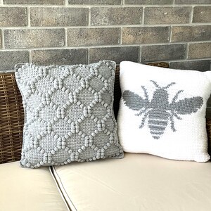May include: Two crocheted throw pillows, one gray with a textured pattern and one white with a gray bee design.