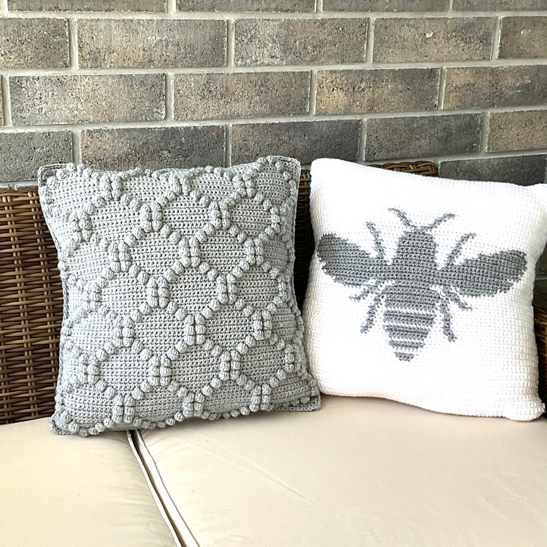 May include: Two crocheted throw pillows, one gray with a textured pattern and one white with a gray bee design.