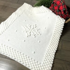 May include: White crocheted baby blanket with a snowflake design. The blanket has a textured border with raised bumps.