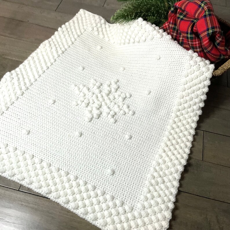 May include: White crocheted baby blanket with a snowflake design. The blanket has a textured border with raised bumps.